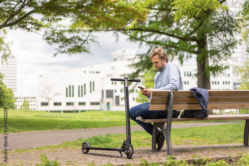 Businessman with E-Scooter sitting on bench in city park using smartphone, Essen, Germany Businessman with E-Scooter sitting on bench in city park using smartphone, Essen, Germany