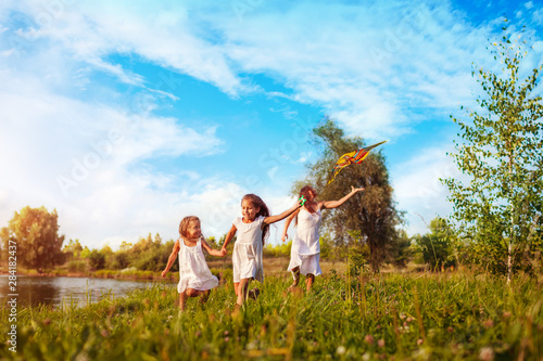 Happy little girls running with kite in summer park while thir mother helps them. Children having fun playing outdoors photo