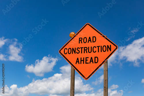closeup of road construction ahead sign and warning light with blue sky and clouds in background
