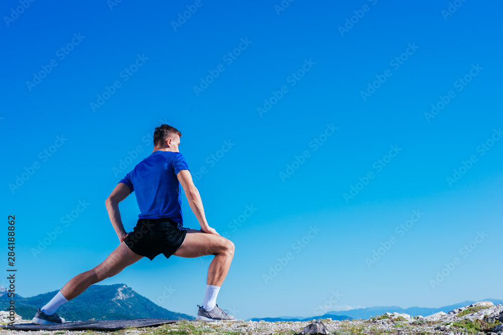 Blonde athlete wearing blue dry-fit shirt stretching and preparing for a workout while standing on top of a cliff which has an amazing view. Blonde athlete wearing blue dry-fit shirt stretching and preparing for a workout while standing on top of a cliff which has an amazing view.