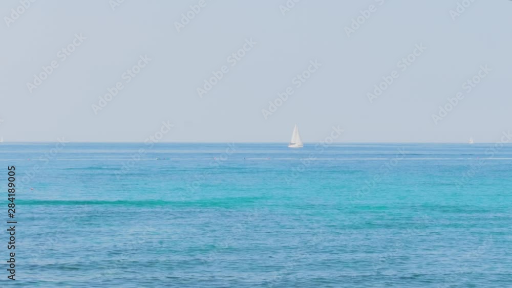 White sail on aquamarine sea, unrecognizable swimming people, oncoming sea waves against clear blue sky. Handheld. Waves washing seaside, swimmers, yacht. Collapse of shore incoming waves, sailboat White sail on aquamarine sea, unrecognizable swimming people, oncoming sea waves against clear blue sky. Handheld. Waves washing seaside, swimmers, yacht. Collapse of shore incoming waves, sailboat