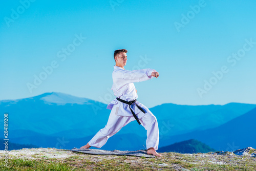 Karate man in a kimono performs a front hand kick (Choku-zuki) while standing on the green grass on top of a mountain.