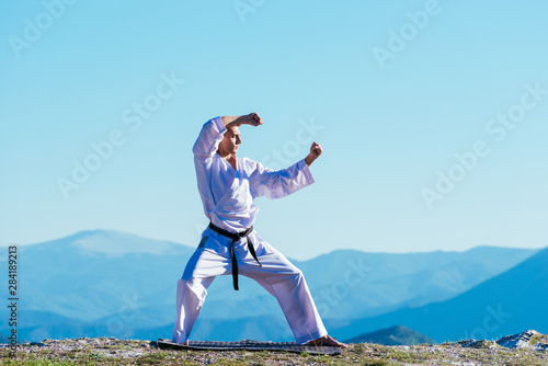 Blond karate athlete does kata on top of a mountain while performing a line up of kicks, punches and blocks on top of a mountain on a sunny day.