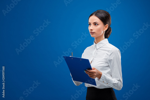 Young businesswoman seriously thinking about an idea while holding a blue clipboard, isolated on blue background.