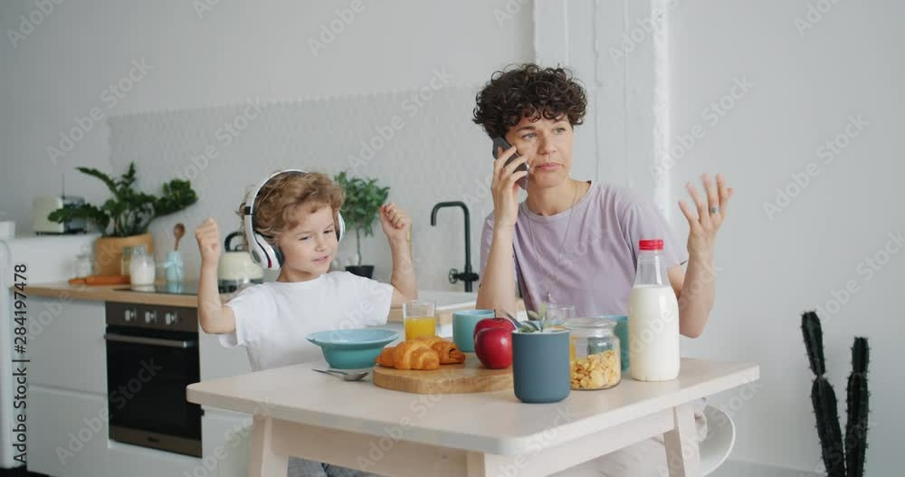 Girl is talking on mobile phone in kitchen at home while small son is listening to music in headphones having fun. Modern devices, lifestyle and apartment concept. Girl is talking on mobile phone in kitchen at home while small son is listening to music in headphones having fun. Modern devices, lifestyle and apartment concept.