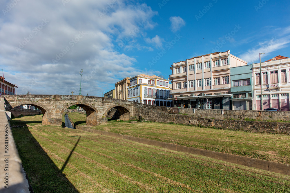 Bridge over river with green grass and old buildings of the city hall of the historic city of Sao Joao del Rey in the state of Minas Gerais, Brazil Bridge over river with green grass and old buildings of the city hall of the historic city of Sao Joao del Rey in the state of Minas Gerais, Brazil
