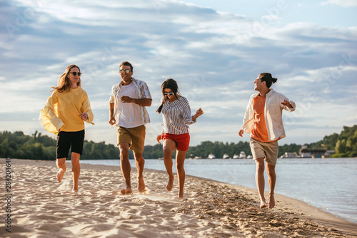 cheerful multicultural friends running on sand beach near river