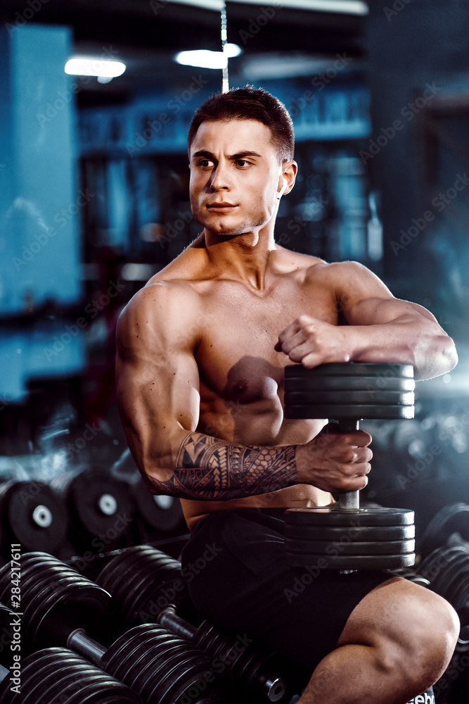 young adult man handsome athlete working out in gym, sitting on a bench and holding dumbbell with raised arms. indoor, looking weight.