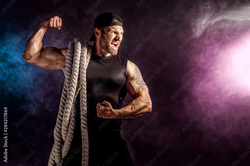 Bearded athletic looking bodybulder holding battle rope on dark studio background with smoke. Strength and motivation.