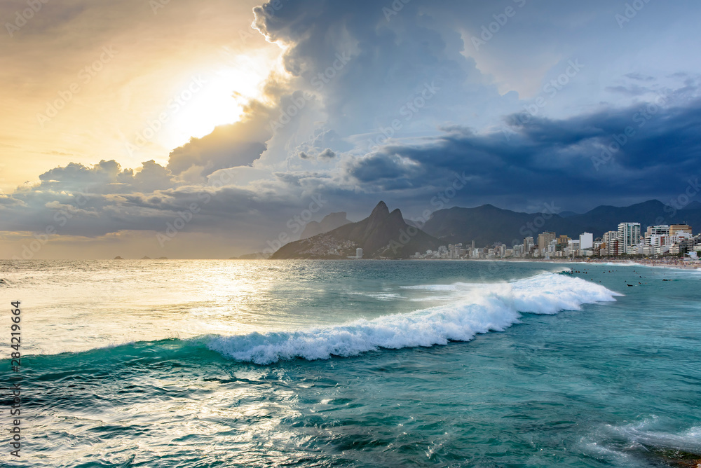 Naklejka premium Summer sunset with cloudy sky, surfers and citi view with Two Brothers hill and Vidigal slum view from Ipanema beach at Rio de Janeiro
