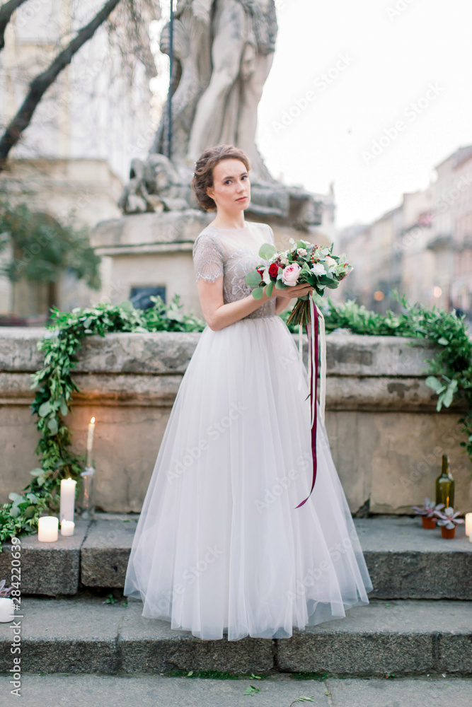 Portrait of beautiful bride in amazing wedding dress holding tender bouquet while standing outdoors in the stairs. Old city center, Lviv, Ukraine