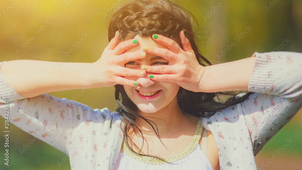 Fototapeta premium Girl put her hands and fingers to his forehead to shield his eyes from the sun, looks up and happy. Portrait of a beautiful brunette girl covering face by hands of bright sun light, summer vacation.