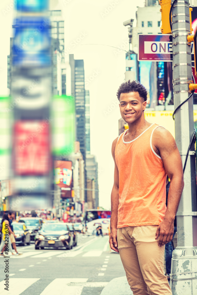 Obraz premium Young Happy Mix Race American Man traveling in New York City in hot summer, wearing orange tank top, beige pants, standing on intersection of street in Times Square of Manhattan, looking, smiling..