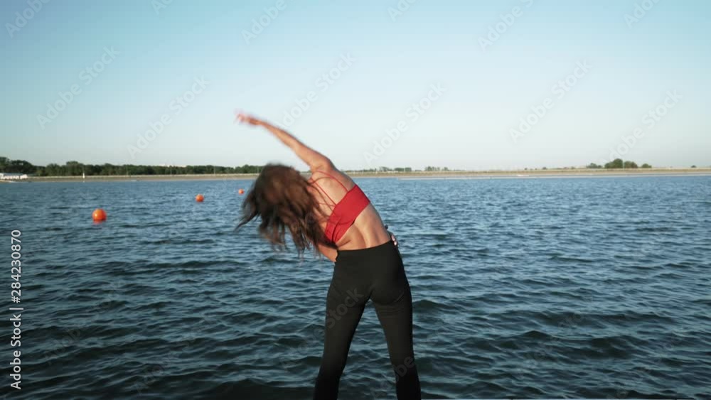 Young athletic Caucasian girl in a red tank top and leggings does warm-up before training against the background of the lake. He is engaged in fitness. Young athletic Caucasian girl in a red tank top and leggings does warm-up before training against the background of the lake. He is engaged in fitness.