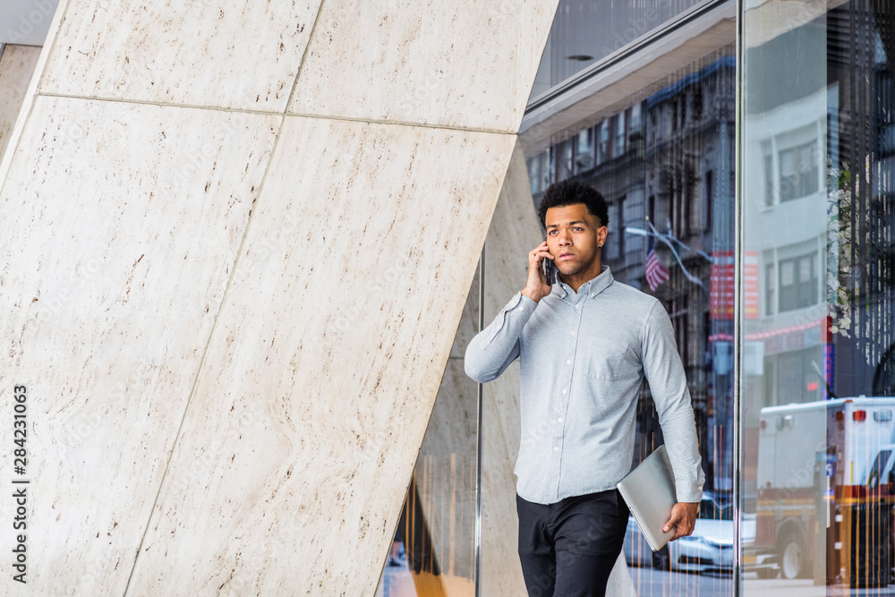 Young Mix Race American Businessman talking on cell phone, traveling, working in New York City, wearing light gray long sleeve shirt, carrying laptop computer, walking to street from office building..