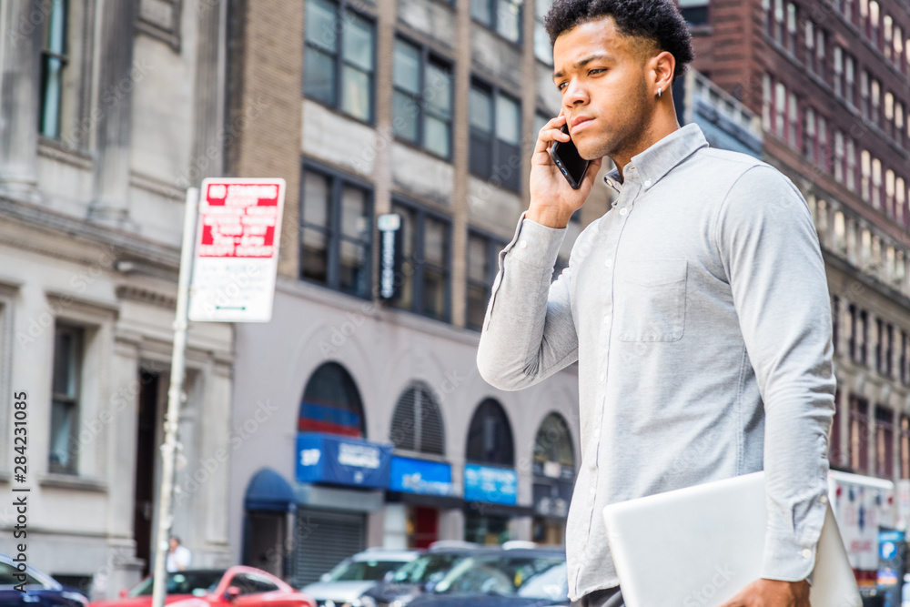 Young Mix Race American Businessman talking on cell phone, traveling in New York City, wearing gray shirt, carrying laptop computer, walking on street in Manhattan. Cars, buildings on background..