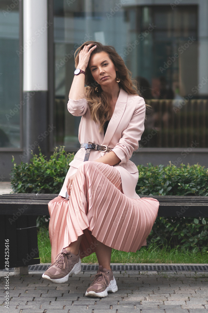 Fototapeta premium girl sitting on a bench on the street near the building with shop windows. Coral, pink jacket, pleated skirt, sneakers. Young beautiful European.