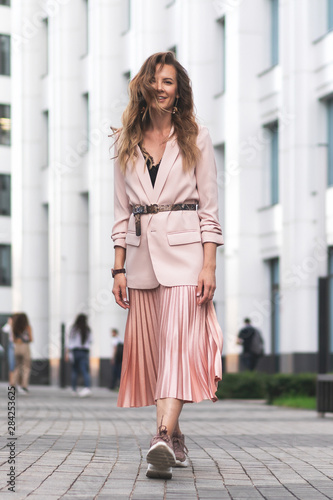 women's jacket and long skirt, pink, peach-colored pleated. The girl walks on the modern stone pavement. Long wavy hair, pretty face. Copy space