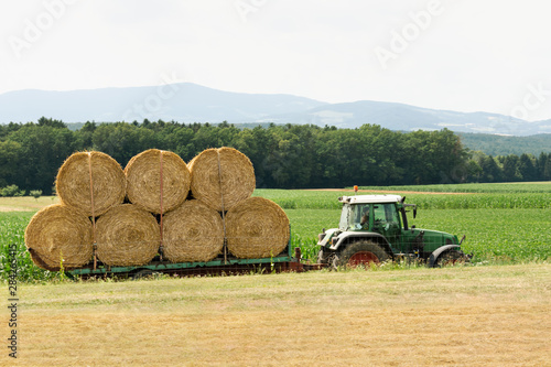 A tractor rides on a road amid fields and carries bales of hay for storage.