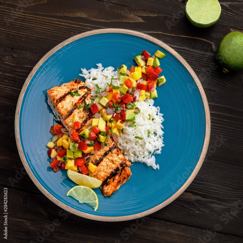 Grilled salmon fillet with fruit sauce made of mango, bell pepper, onion and avocado with basmati rice garnish on a blue plate. Delicious seafood meal. Top view shot above.