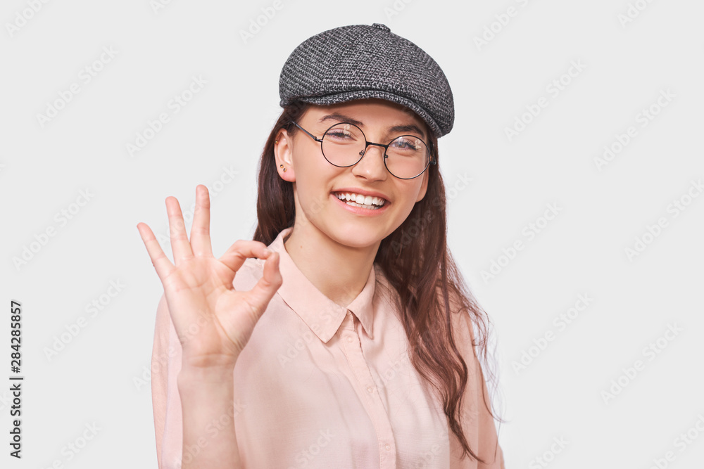 Studio portrait of positive young woman wears trendy gray cap, pink shirt and round transparent eyeglasses, showing Ok gestures. Pretty student girl smiling, blinking with an eye and showing okay sign Studio portrait of positive young woman wears trendy gray cap, pink shirt and round transparent eyeglasses, showing Ok gestures. Pretty student girl smiling, blinking with an eye and showing okay sign