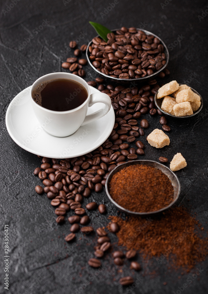 White cup of fresh raw organic coffee with beans and ground powder with cane sugar cubes with coffee tree leaf on black background. Top view