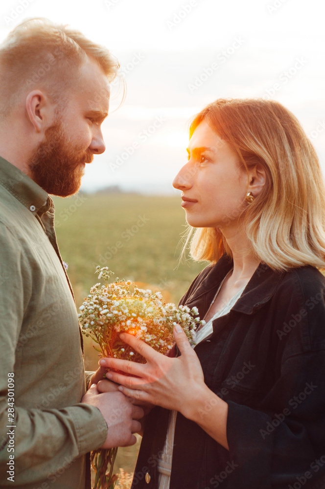 Handsome guy and blonde girl walking on the field on a beautiful warm sunset.  standing in the sun. bouquet of field flowers