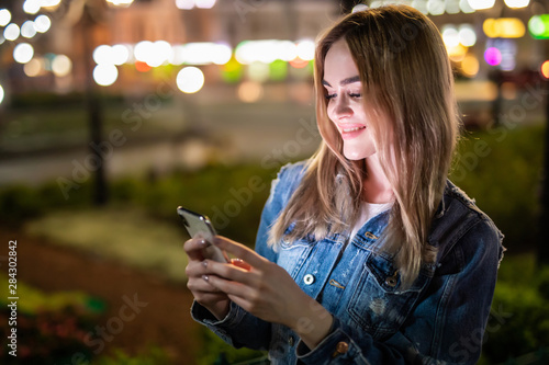Young woman holding a phone in hands texting at night with city lights on background