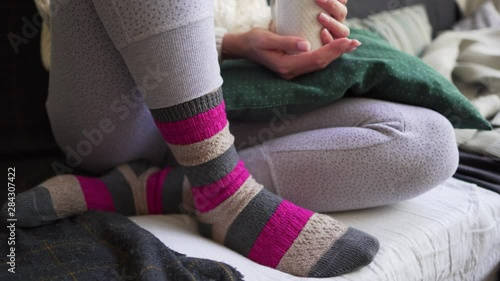 Pink and brown warm woolen socks close-up - a woman is sitting with a white cup of hot drink on the sofa with a plaid and pillows. Lagom or Hygge concept - comfort and simplicity