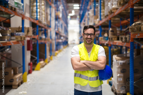 Portrait of middle aged warehouse worker standing in large warehouse distribution center with arms crossed. In background shelves with goods.