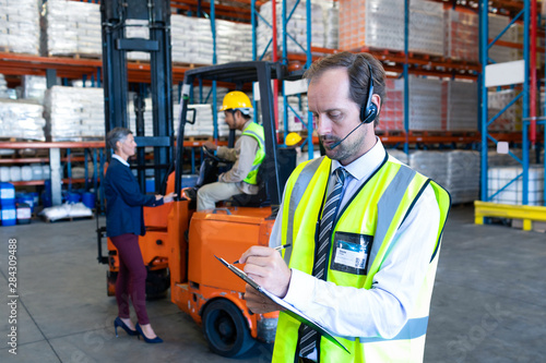 Male supervisor with headset writing on clipboard in warehouse