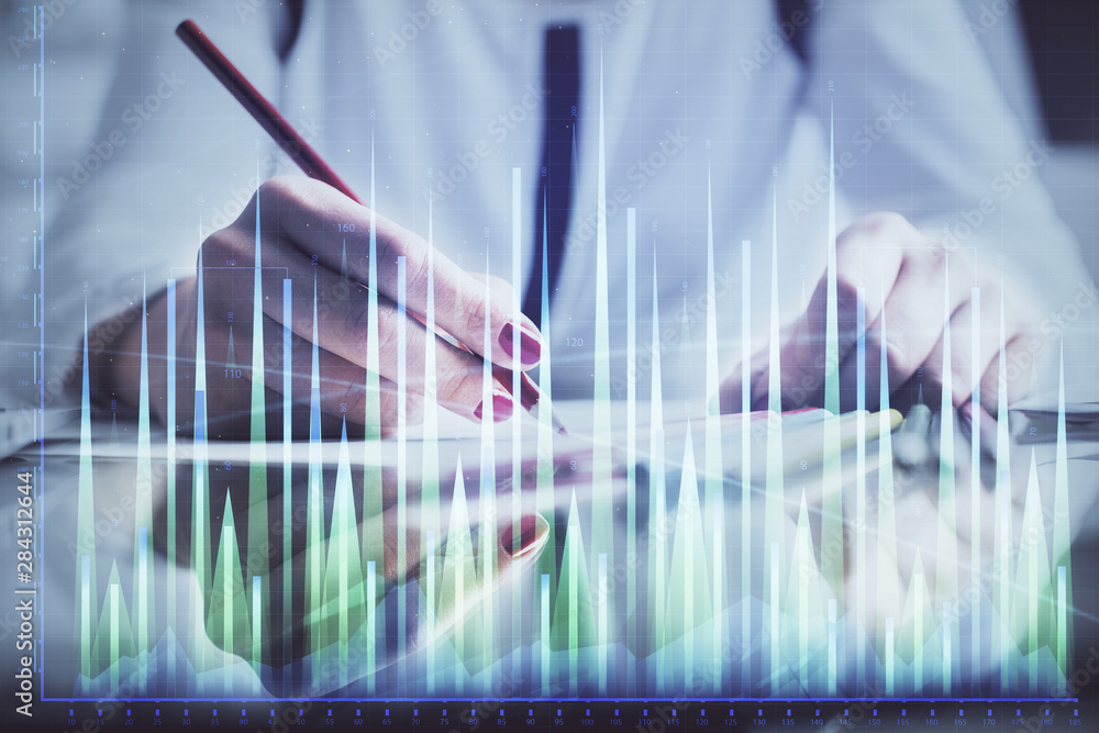 A woman hands writing information about stock market in notepad. Forex chart holograms in front. Concept of research. A woman hands writing information about stock market in notepad. Forex chart holograms in front. Concept of research.