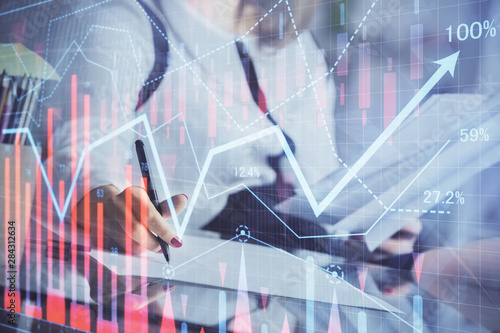 A woman hands writing information about stock market in notepad. Forex chart holograms in front. Concept of research.