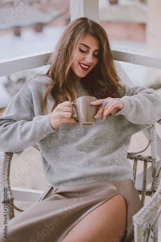 Relaxed woman with cup of coffee or tea. Stylish girl dressed in a gray sweater and silk skirt resting on the balcony.