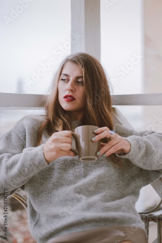 Relaxed woman with cup of coffee or tea. Stylish girl dressed in a gray sweater and silk skirt resting on the balcony.