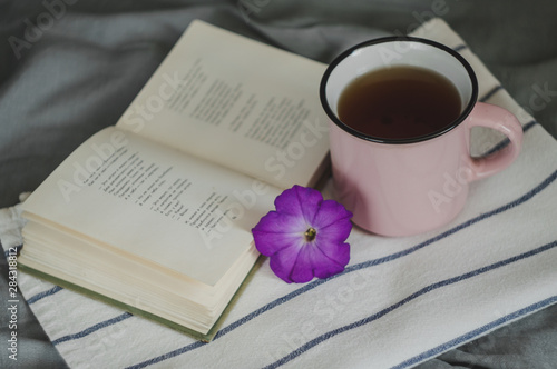 Cup of tea, old book and purple flower on white striped towel, on gray background close up