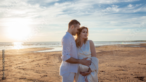 Love, wait for the baby. Portrait of a beautiful couple in white kissing on the background of the sunset sea.