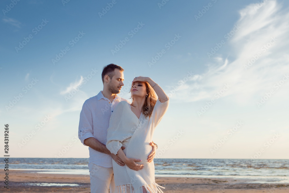 Romantic walk on the beach, waiting for a baby. Happy young couple hugging tummy, spending time by the sea.