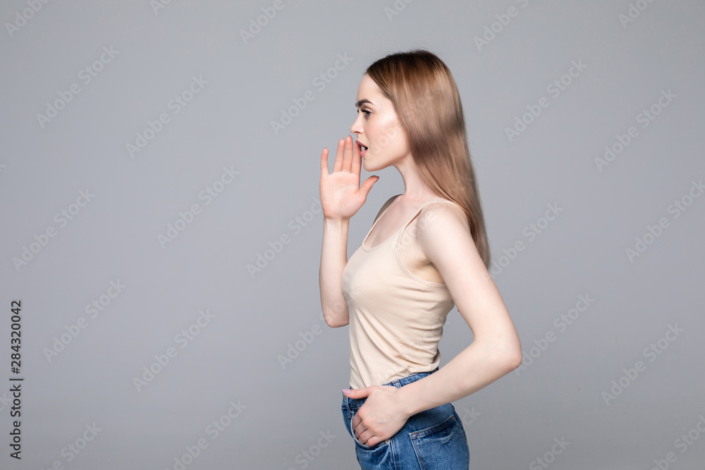 Side-view profile portrait of attractive woman telling news, shouting and holding hand near her open mouth isolated on gray background Side-view profile portrait of attractive woman telling news, shouting and holding hand near her open mouth isolated on gray background