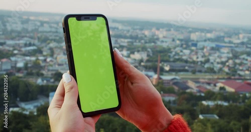 Close-up female hands holding green chromekey mobile phone mock-up. Excited girl using smartphone while staying in mountain park observing modern city panorama. photo