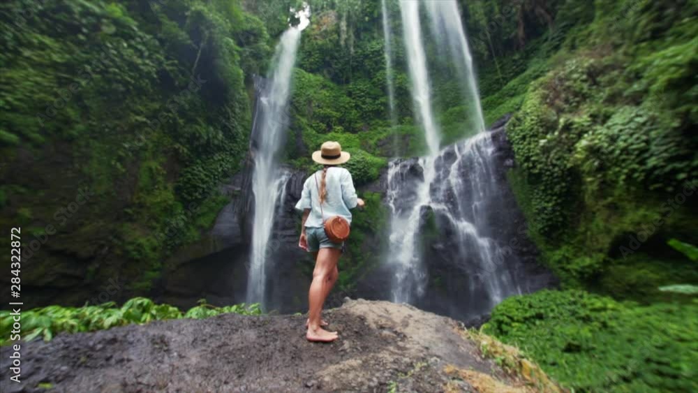 Slow motion view from below of barefooted woman standing on big stone and enjoying amazing high waterfall Sekumpul in Bali rainforest. Great wild nature of Indonesia Slow motion view from below of barefooted woman standing on big stone and enjoying amazing high waterfall Sekumpul in Bali rainforest. Great wild nature of Indonesia