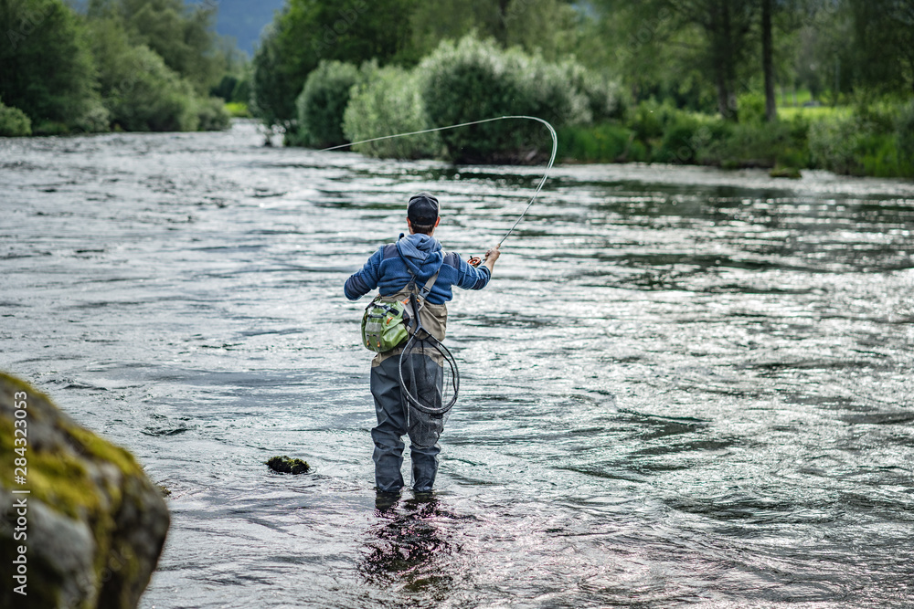Fliegenfischer in Wathose mit Angelroute im Fluss stehend vor Wald und Sträuchern Fliegenfischer in Wathose mit Angelroute im Fluss stehend vor Wald und Sträuchern