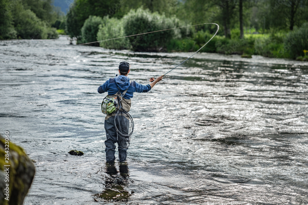 Fliegenfischer in Wathose mit Angelroute beim Werfen im Fluss vor Bäumen und Steinen Fliegenfischer in Wathose mit Angelroute beim Werfen im Fluss vor Bäumen und Steinen