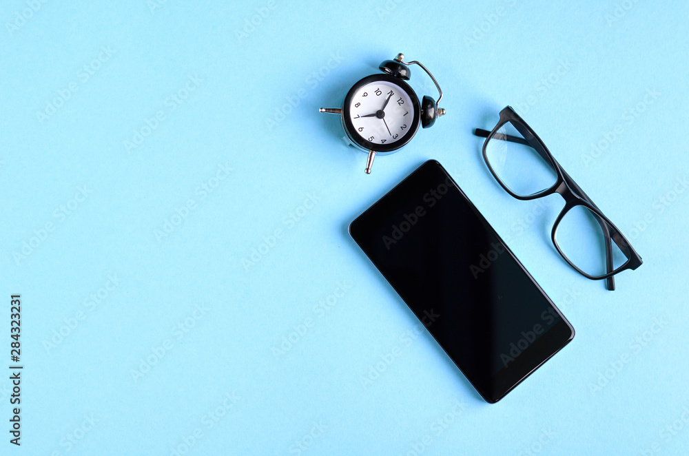 Black glasses, alarm clock and cellphone on blue background composition. Black glasses, alarm clock and cellphone on blue background composition.