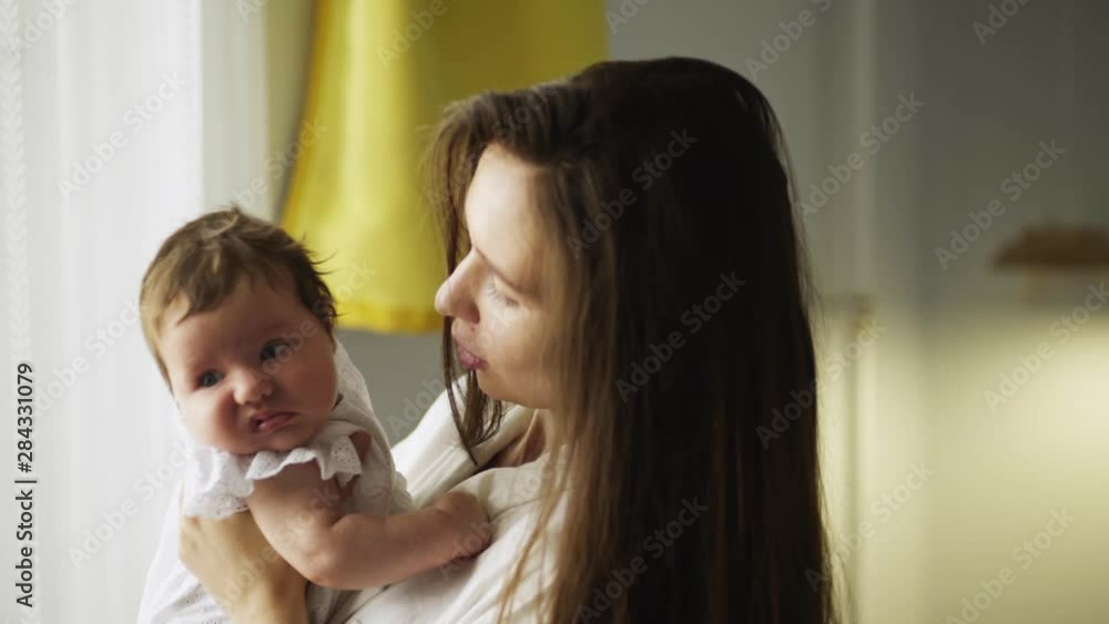 Young mother with her newborn child on hands standing near the window. Side view pretty female nursing baby. Woman and new born girl enjoying togetherness in a bedroom. Family at home Young mother with her newborn child on hands standing near the window. Side view pretty female nursing baby. Woman and new born girl enjoying togetherness in a bedroom. Family at home