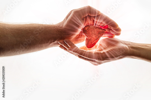 Female and male hands are keeping red heart together. Heart with brightly ray of sunshine is between hands. World Kindness Day.