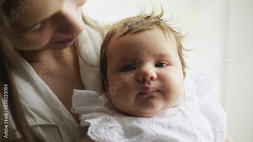 Young mother holding her newborn child near the window. Close-up portrait newborn baby nursing on hands. Woman and new born girl relax in a bedroom. Family at home photo