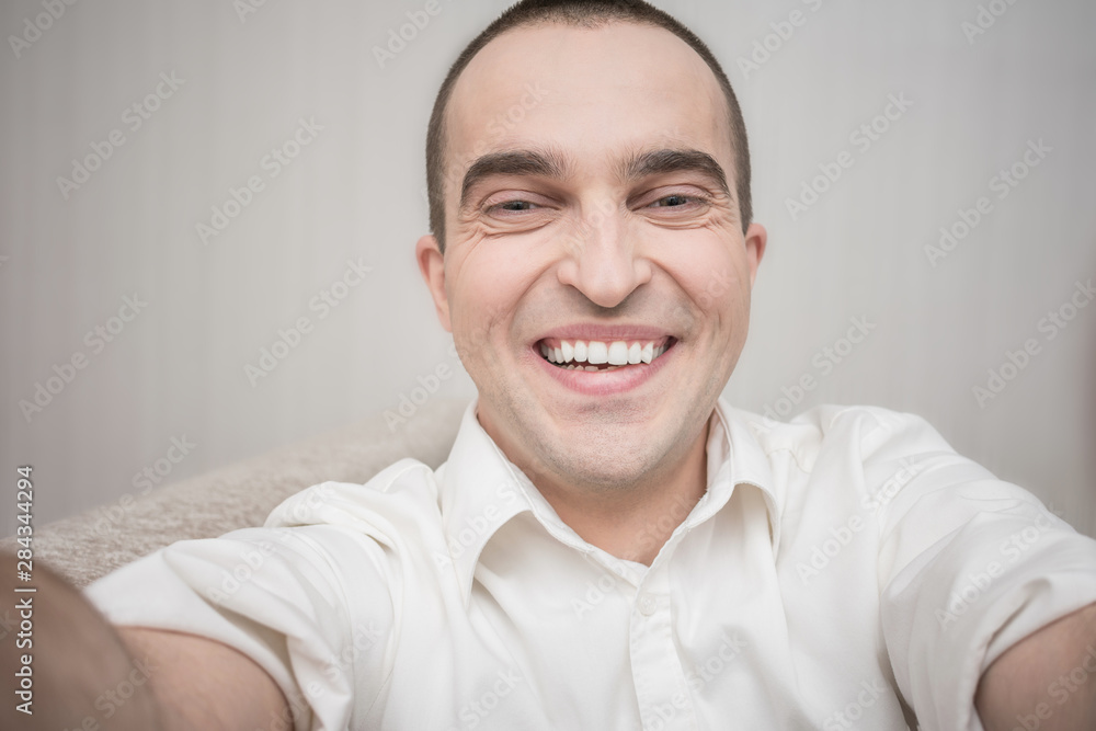 Attractive young man in white shirt laughing for selfie, sitting at home on the couch, front view, toned Attractive young man in white shirt laughing for selfie, sitting at home on the couch, front view, toned