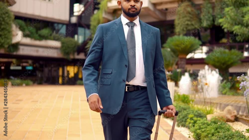 Indian businessman wearing blue suit walking with travel bag or suitcase near office or hotel. Travel business concept Indian businessman wearing blue suit walking with travel bag or suitcase near office or hotel. Travel business concept