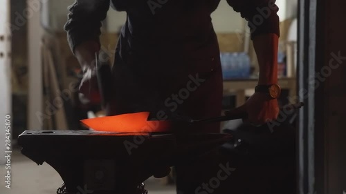 Medium close-up shot of a knifemaker forging a hot orange glowing knife with a small hammer on an old anvil outside in the night with workshop in background - 4K - Real-Time 23.976fps photo